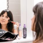 A woman applying makeup using a brush in front of a bathroom mirror, surrounded by cosmetics.